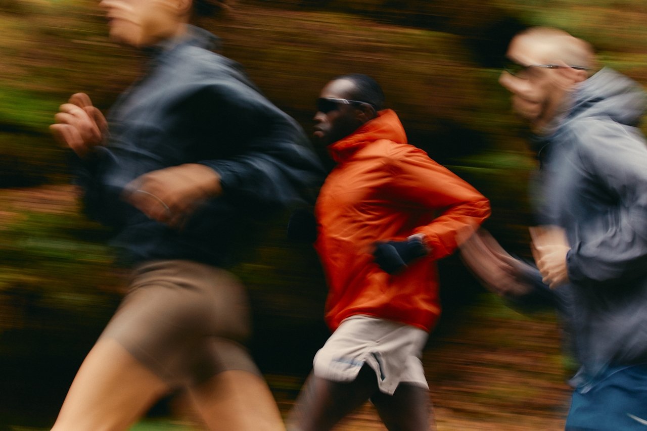 Three runners moving fast along a wooded trail, the center runner in an orange jacket